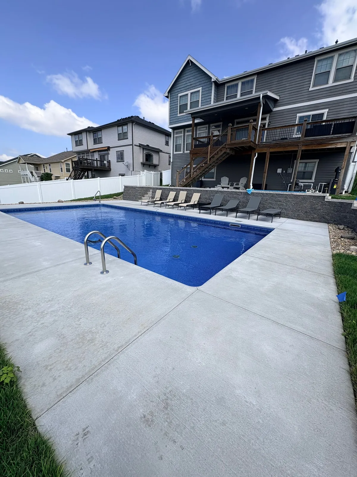 Rectangular vinyl liner inground pool with a ladder, a stone retaining wall and lounge chairs below a gray two-story house, built by Olympic Pool & Spa in the Omaha metro
