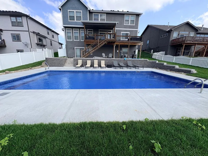 Rectangular inground pool with a broad concrete deck, a stone retaining wall and lounge chairs below a gray two-story house in the Omaha metro, built by Olympic Pool & Spa