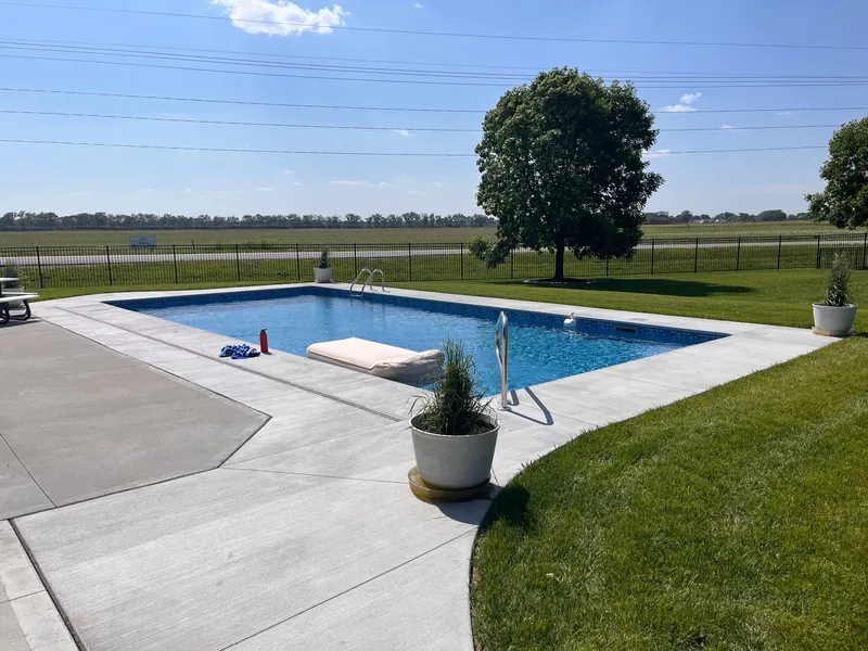 Rectangular inground pool with a tanning ledge, a white concrete deck and a black aluminum fence on an acreage with a farmland view, built by Olympic Pool & Spa