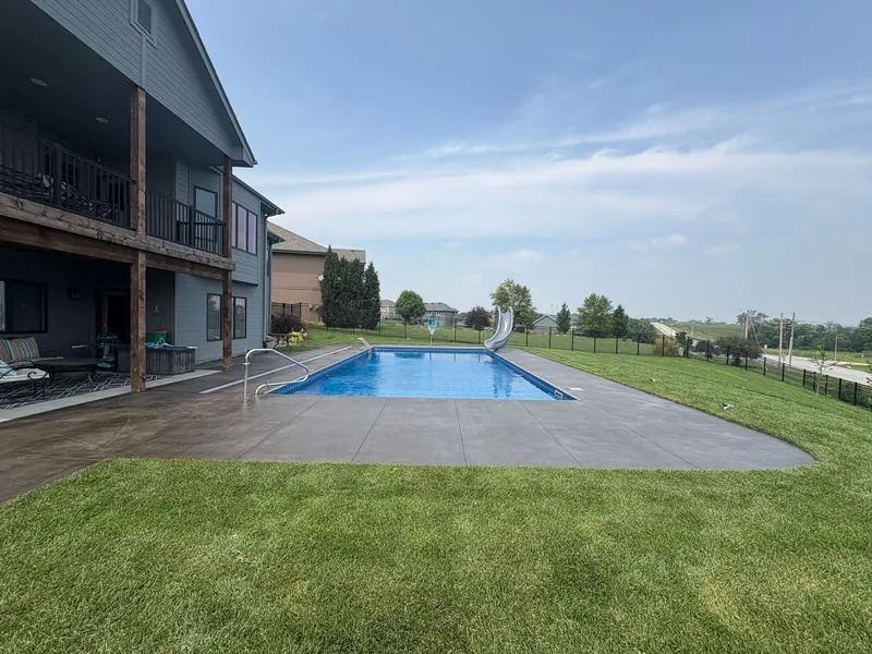 Rectangular inground pool with a slide and a dark stained concrete deck beside a two-story house with a covered patio, built by Olympic Pool & Spa in the Omaha metro